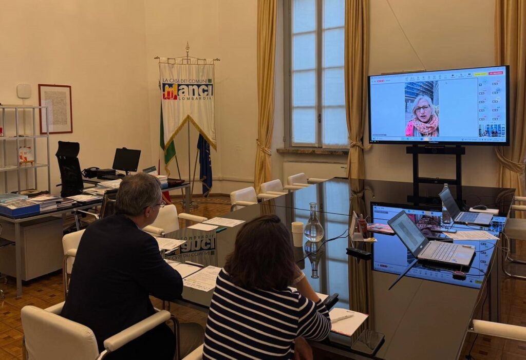 La foto scattata nella sede di ANCI Lombardia, inquadra due relatori che guardano lo schermo durante il webinar